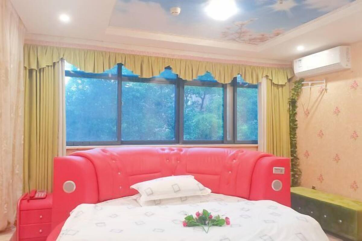 Un dormitorio con un ventanal grande, un cabecero tapizado en rojo, una mesita de noche y una cama con colcha blanca.