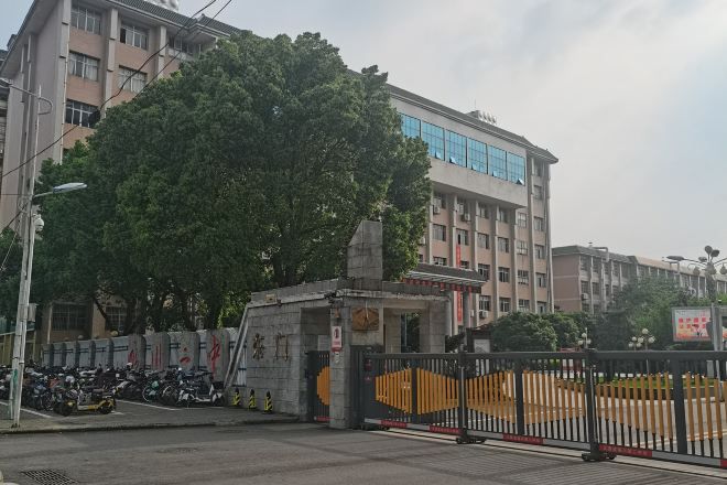 Une entrée fermée par un portail, avec un grand arbre au premier plan, un bâtiment doté de nombreuses fenêtres en arrière-plan et une zone délimitée par une clôture où sont garées des motos.