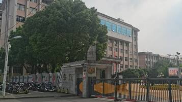 Une entrée fermée par un portail, avec un grand arbre au premier plan, un bâtiment doté de nombreuses fenêtres en arrière-plan et une zone délimitée par une clôture où sont garées des motos.
