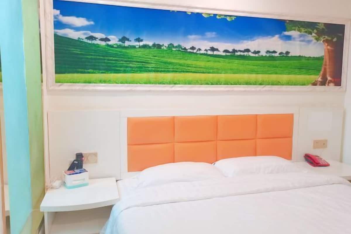 Una habitación de hotel con un ventanal grande, una cama con ropa blanca, un cabecero naranja y una mesita de noche pequeña con un teléfono rojo y una caja de pañuelos.