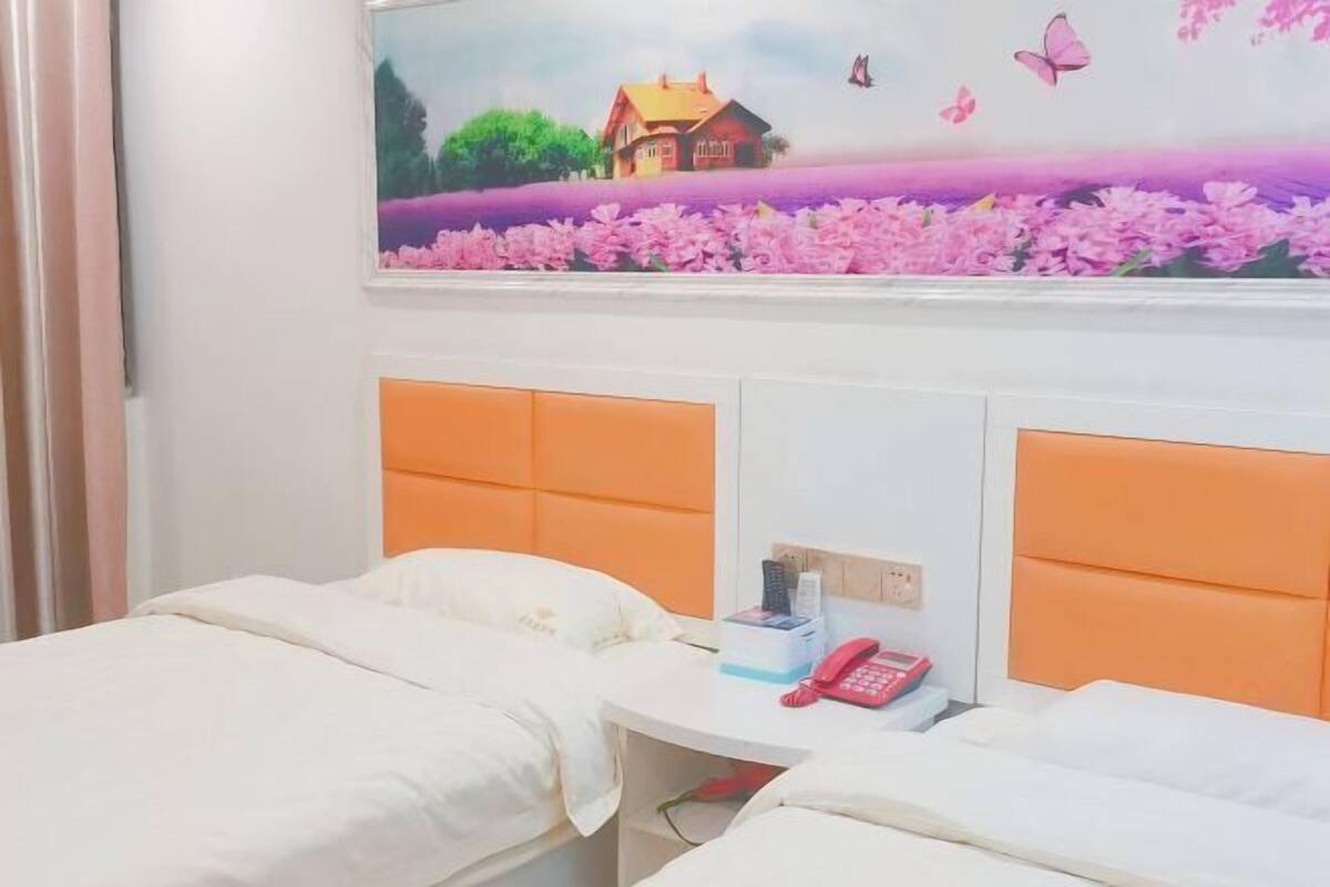 Habitación de hotel con dos camas, cabecero blanco, una mesita con un teléfono y una caja, y un cuadro de una casa y flores en la pared.