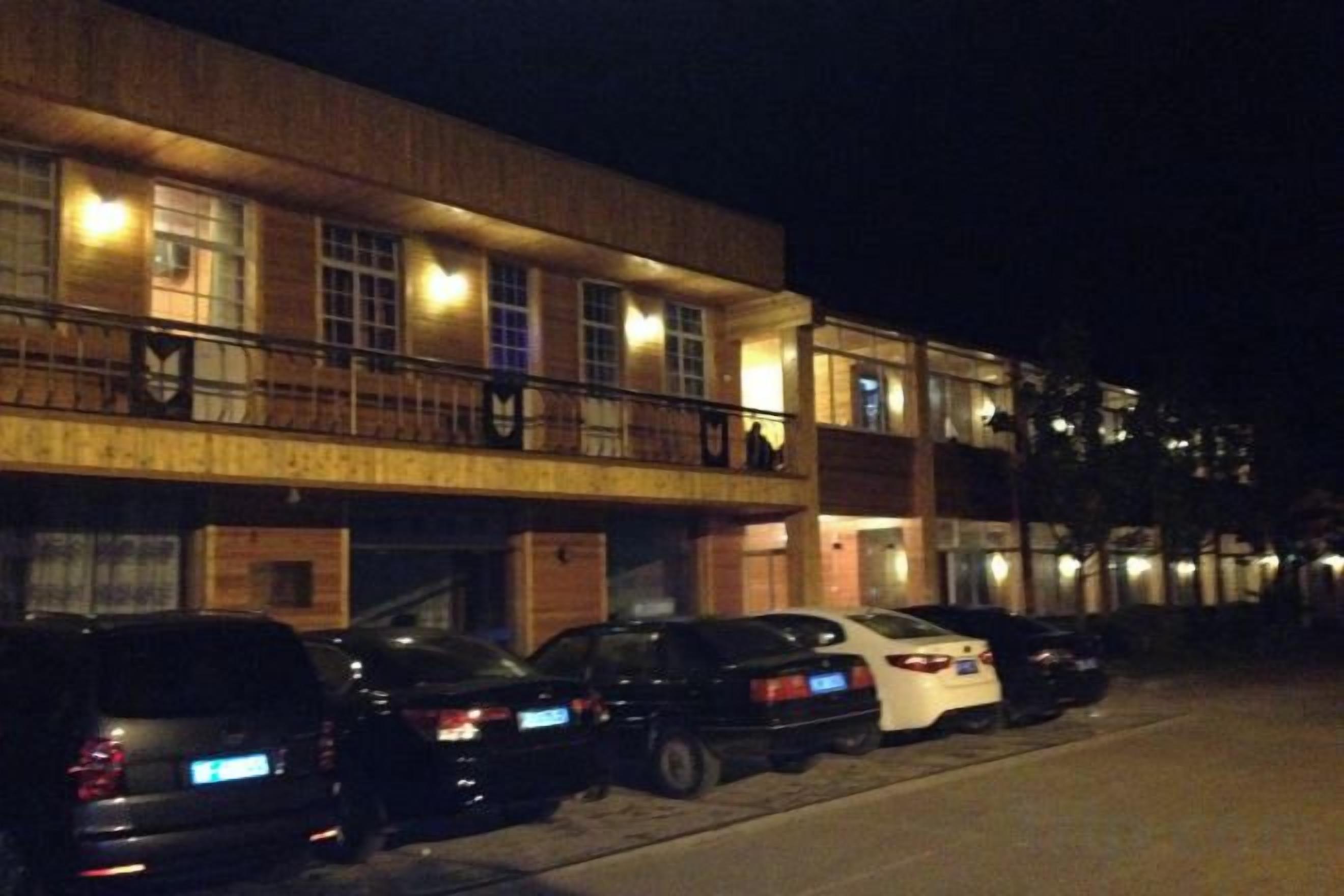 A building with multiple floors, a parking area with several cars, and a balcony with people.