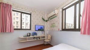 A hotel room with a bed, a desk with a computer, a chair, a window with curtains, and an air conditioning unit. - Chunxin Hostel (Shanghai)