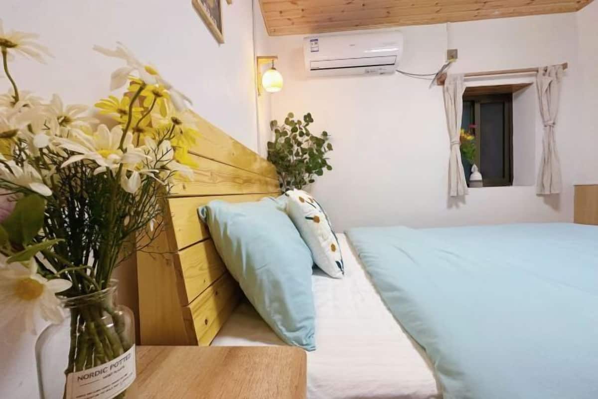 Camera da letto con una testiera in legno, una finestra con tende, un comodino con un vaso di fiori e un letto con biancheria azzurra.