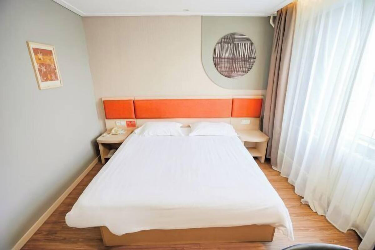 A hotel room with a large bed, a nightstand, a wall art piece, and a window with curtains.