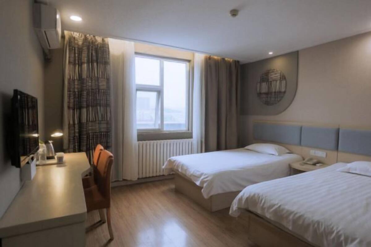 A hotel room with two beds, a desk, a chair, a radiator, and a window with curtains.