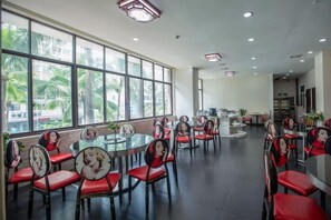 Un comedor con mesas y sillas redondas, ventanas amplias y una cocina al fondo.