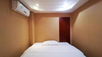 A single bed with a white mattress and pillow in a room with beige walls and a wooden door.