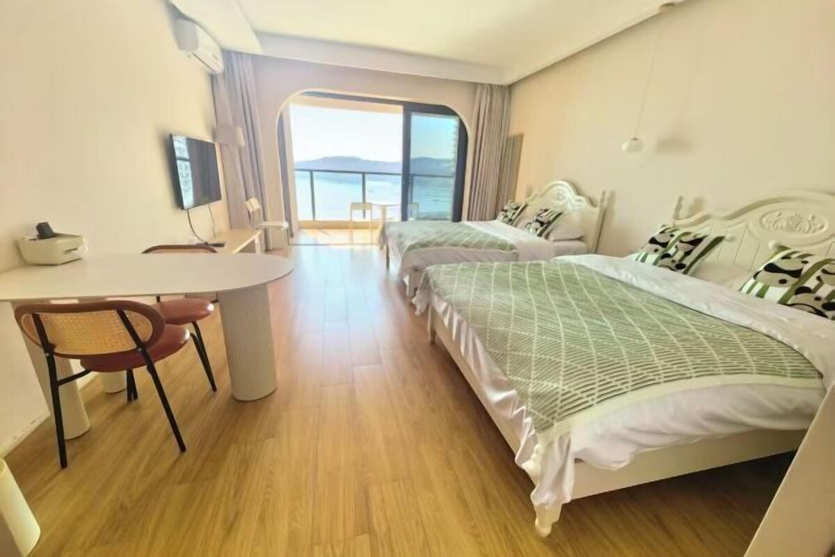 A hotel room with two beds, a desk, and a chair. There is a large window with a view of the sea.