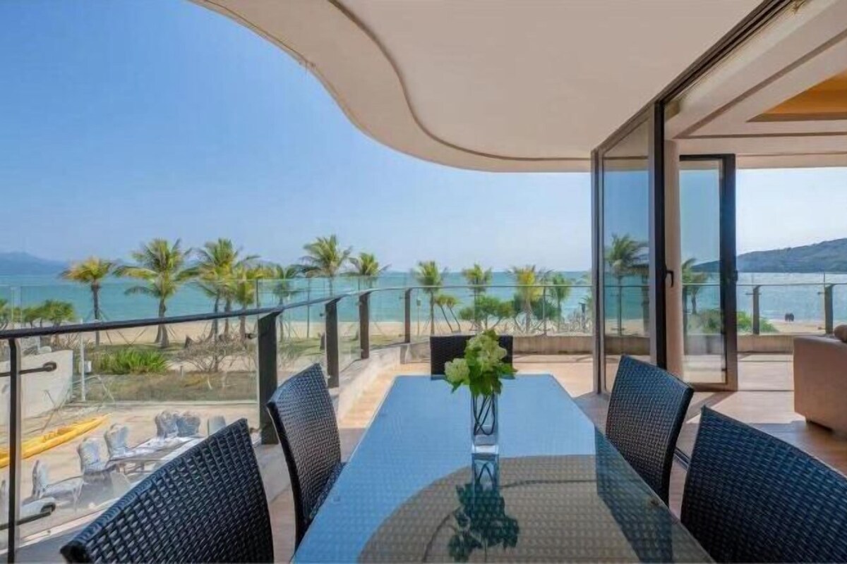 A balcony with a glass table and chairs, overlooking a beach with palm trees and the sea.