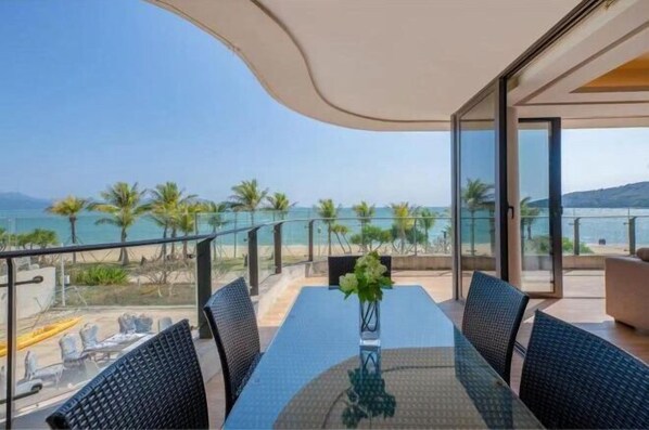A balcony with a glass table and chairs, overlooking a beach with palm trees and the sea. - Relais Boutique Apartment (Huizhou Huarun Xiaojingwan) (Huizhou)