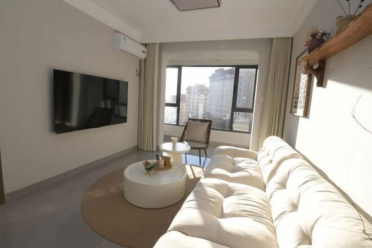 A modern living room with a large flat-screen TV, a round coffee table, a sofa, and a view of tall buildings through large windows.