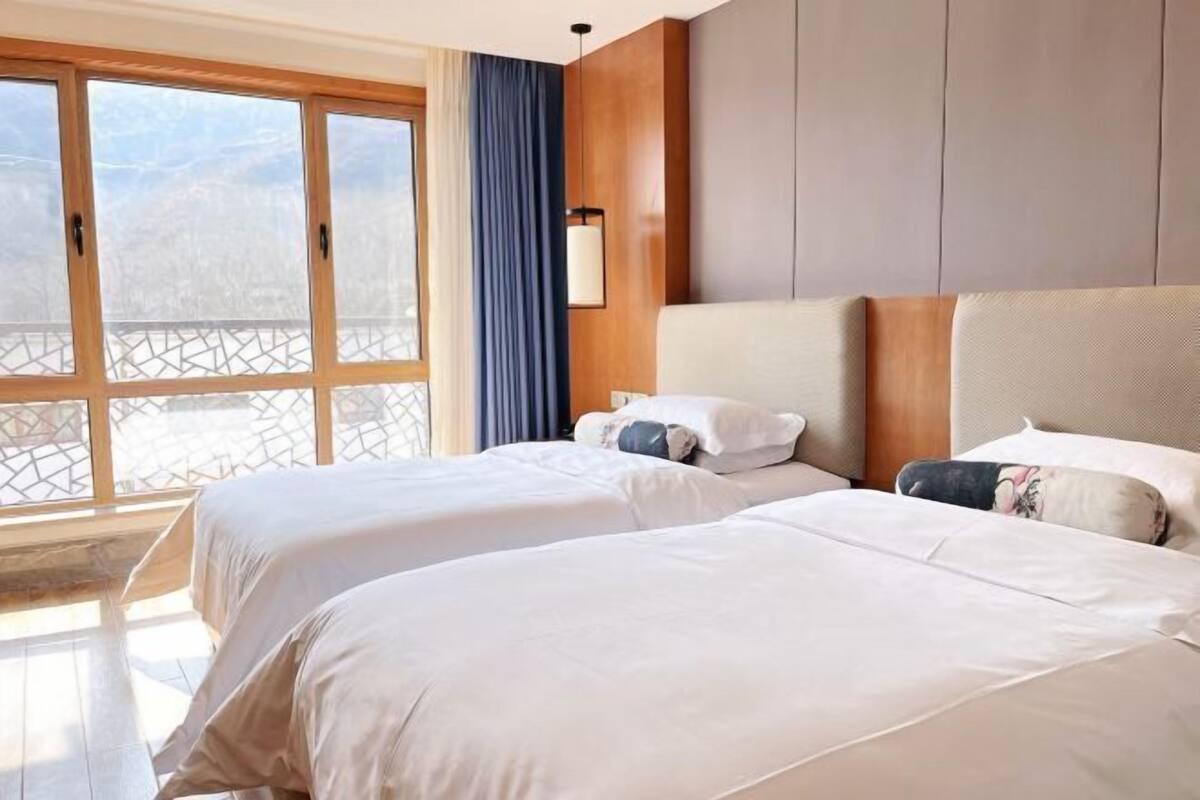 A hotel room with two beds, a large window with a view, a wooden headboard, and a wall-mounted lamp.