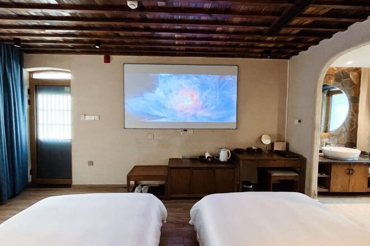 A room with two beds, a TV mounted on the wall, a wooden desk, a bathroom with a stone wall, and a door with curtains.
