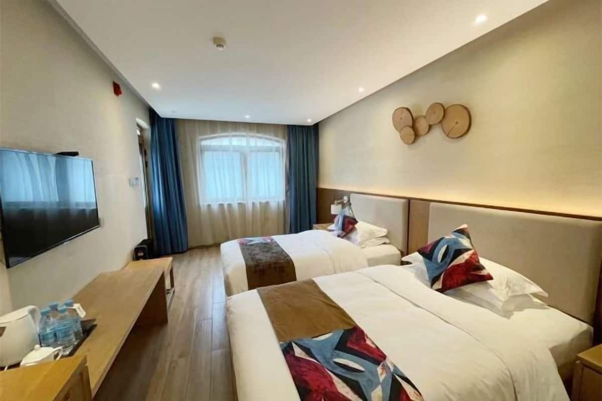 A hotel room with two beds, a flat-screen TV, a wooden bench, and a large window with curtains.