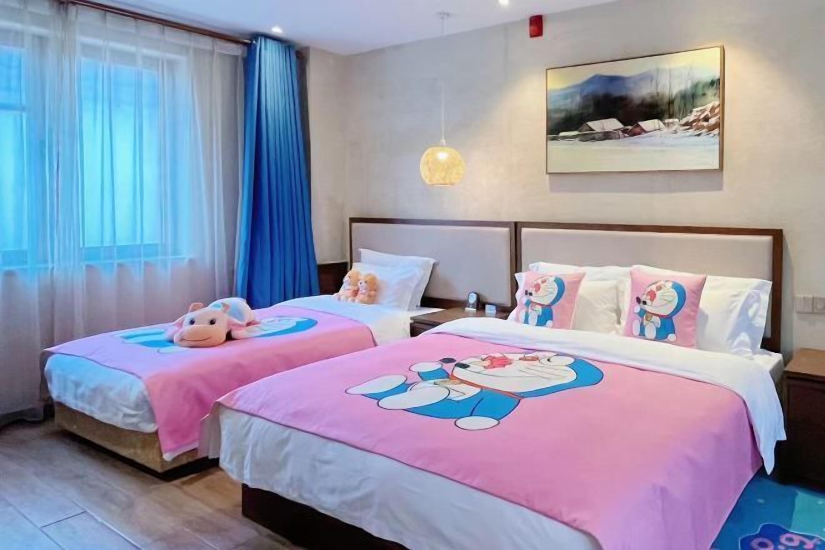 A hotel room with two beds, each with a pink bedspread featuring cartoon characters, a painting on the wall, and a window with sheer curtains.