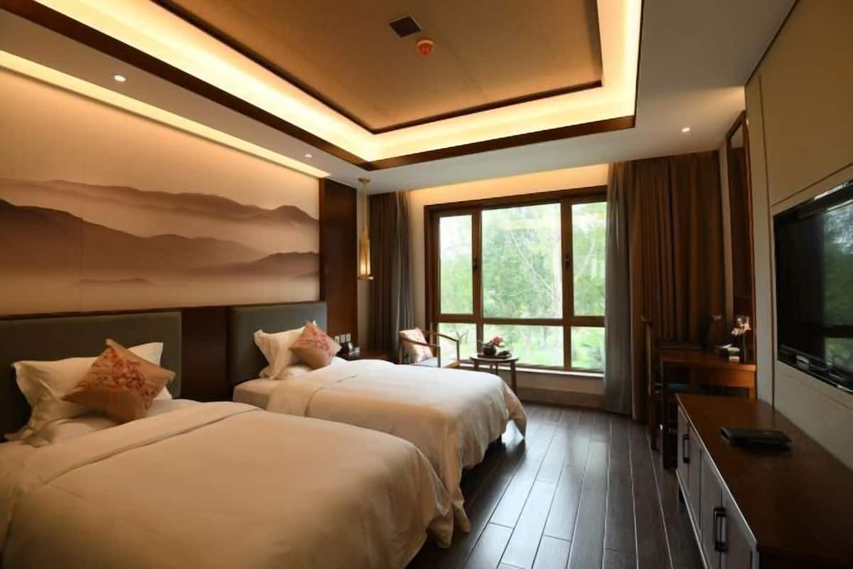 A hotel room with two beds, a TV, a desk, and a large window with a view of greenery.