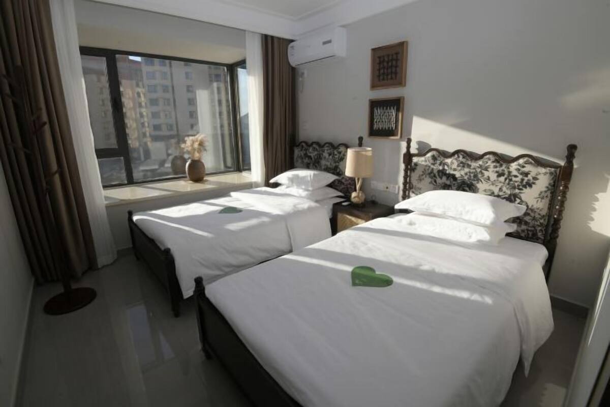 A hotel room with two beds, a window with curtains, a bedside table with a lamp, and a wall-mounted air conditioner.