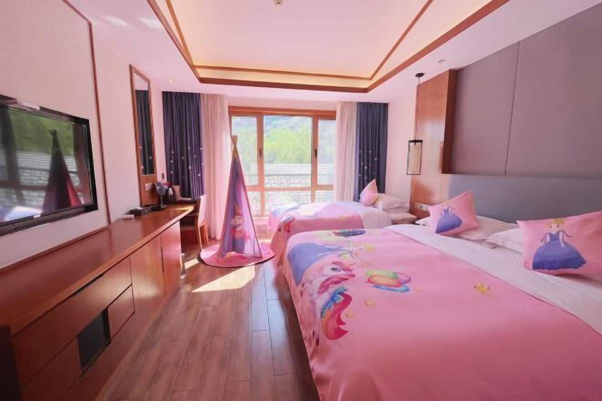 A hotel room with a large TV, a desk, a bed with pink bedding, and a window with curtains.