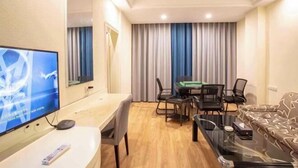 Habitación de hotel con televisión de pantalla plana, una mesa de comedor con sillas, un sofá y un ventanal con cortinas.