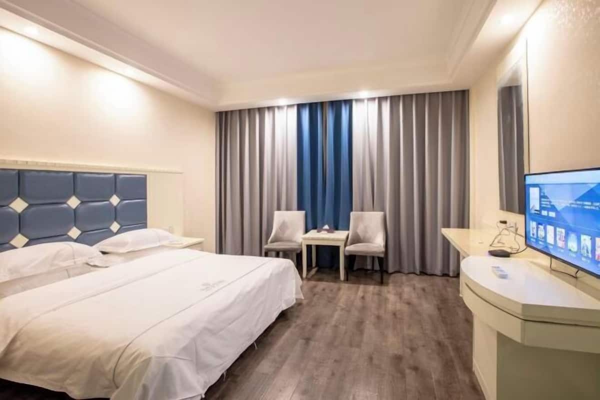 A hotel room with a bed, a desk, two chairs, a TV, and a large window with curtains.