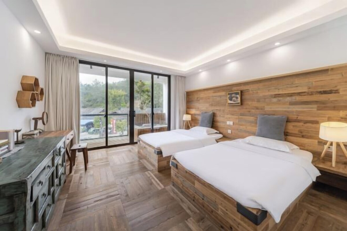 Habitación de hotel moderna con dos camas, cabecero de madera, escritorio con cajones y un amplio ventanal corredero que da a un espacio al aire libre.