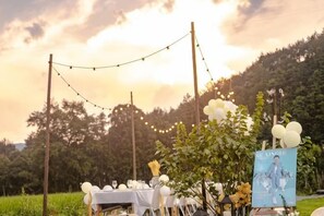 Un área de comedor al aire libre con una mesa dispuesta para una comida, rodeada de follaje y un atardecer de fondo.