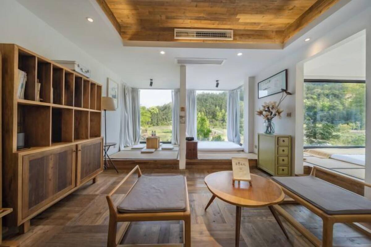 Een moderne kamer met houten meubels, grote ramen en uitzicht op groen.