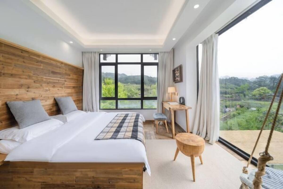 Een slaapkamer met een groot raam, een houten hoofdbord, een bed met wit beddengoed, een bureau en een klein rond tafeltje.