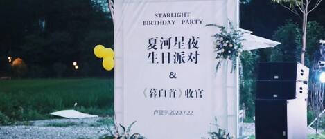 A banner with text for a birthday party, a stage with speakers, and a chair in a grassy outdoor setting.
