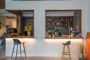 Una zona de bar con mostrador de madera, taburetes y estantería con botellas.