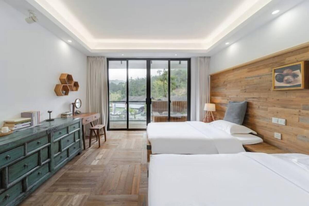 Een hotelkamer met twee bedden, een houten dressoir, een bureau en een groot raam met uitzicht op groen.