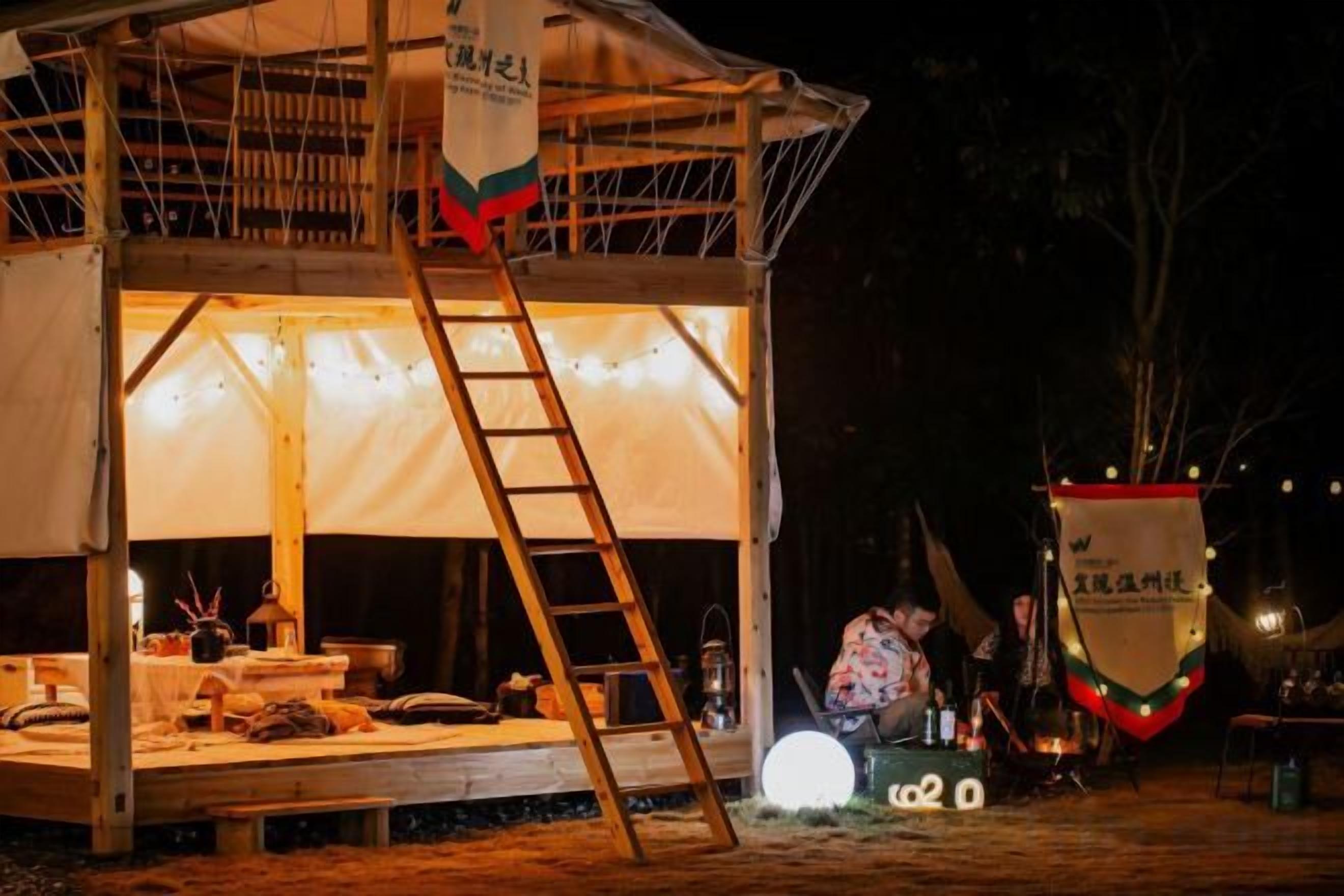Una carpa con una escalera, una mesa con objetos y una persona trabajando cerca de un farol.