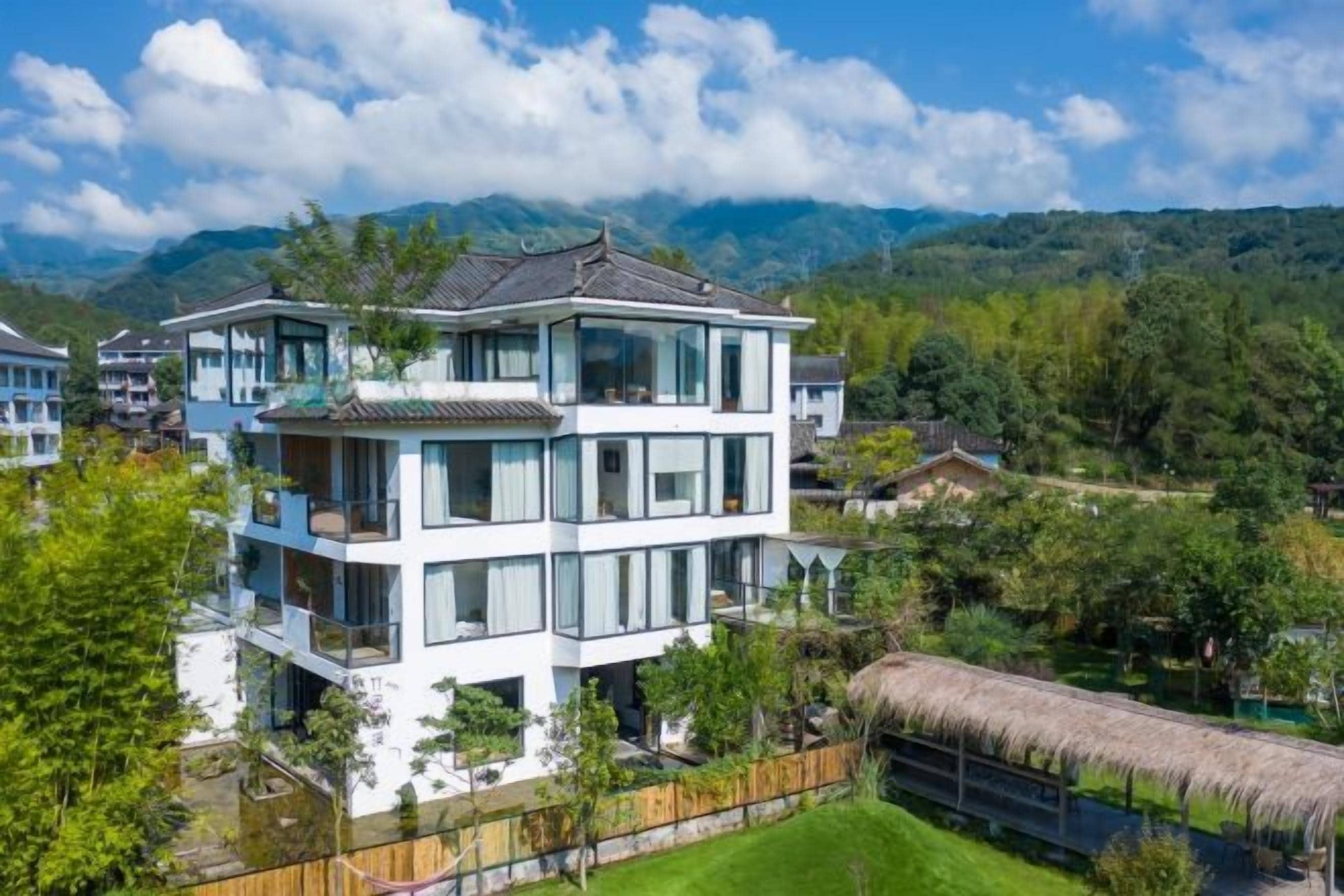 A multi-story building with a balcony, surrounded by greenery and mountains.
