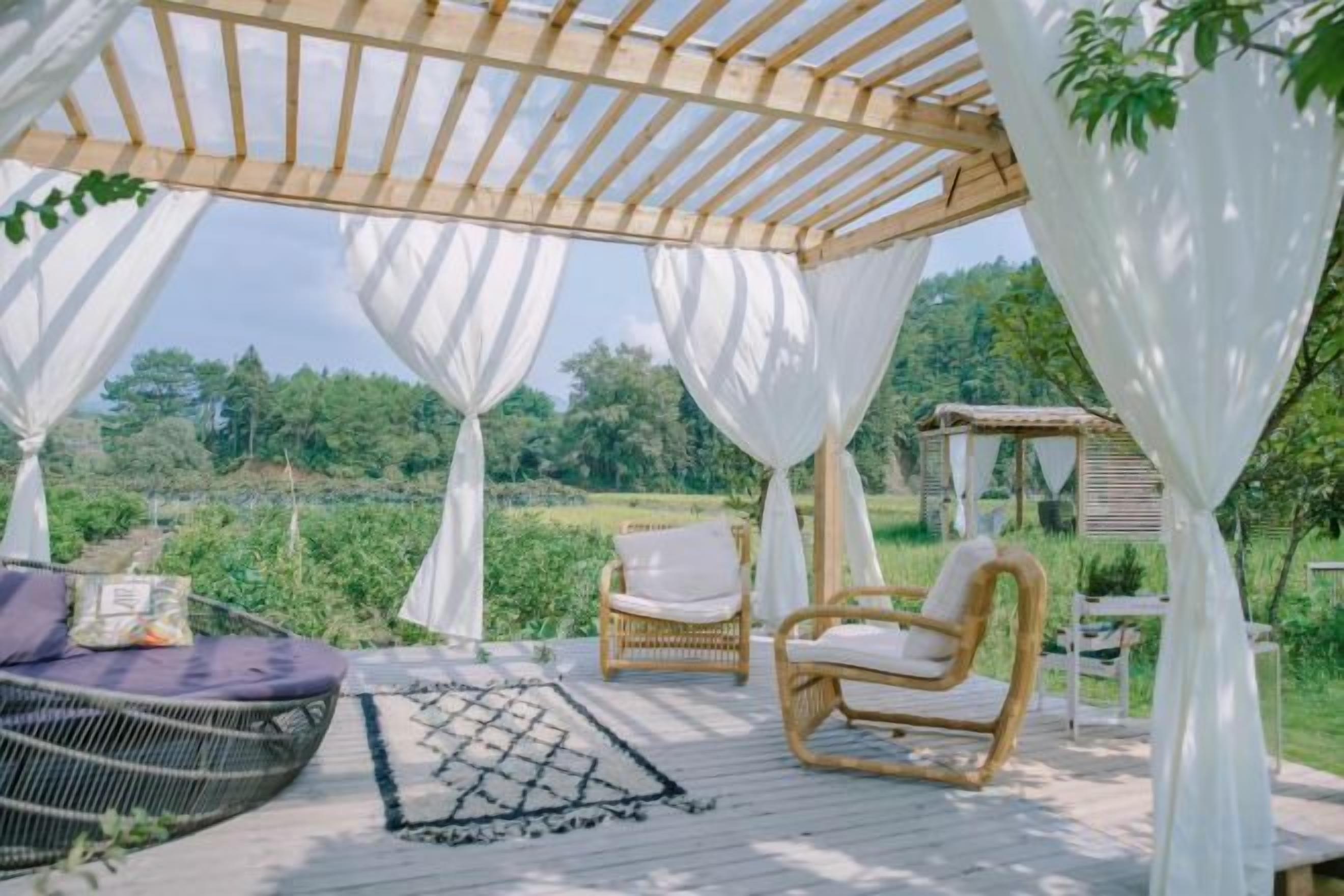 An outdoor patio with a wooden pergola, white drapes, wicker furniture, and a view of a green landscape.