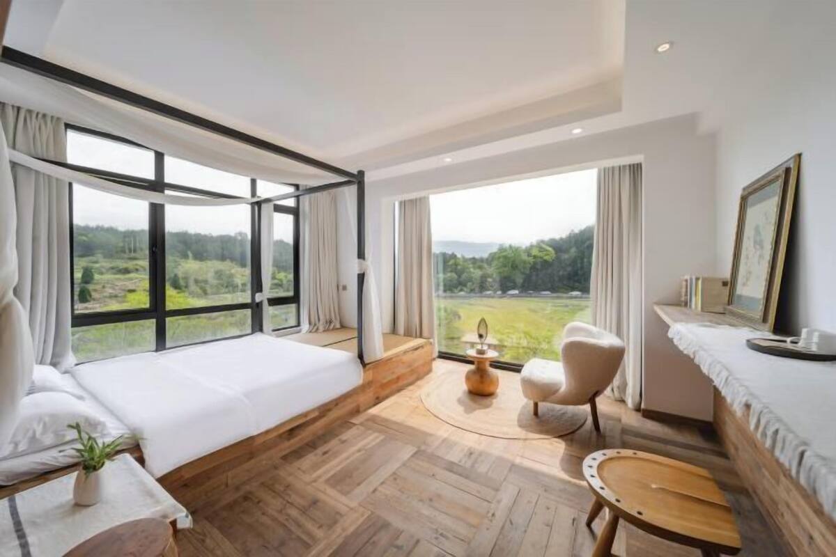 Een moderne slaapkamer met een groot bed, een ronde houten tafel, een stoel en uitzicht op een groen landschap door grote ramen.