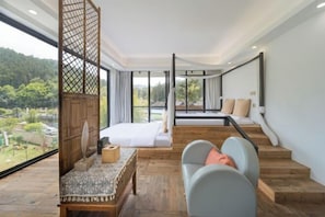 A modern bedroom with a large window, a bed, a seating area, and a view of a forested area.