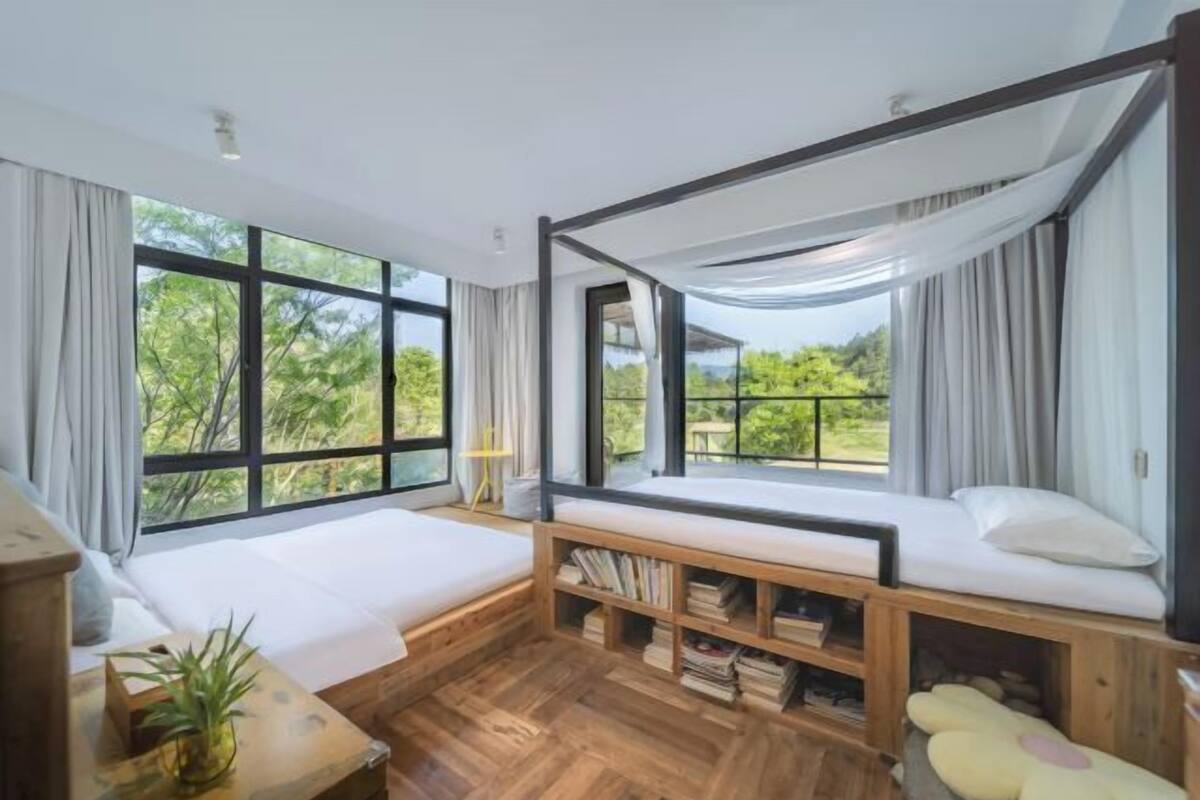 Een kamer met een groot raam, een bed, een houten bankje en een boekenkast.