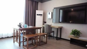 A compact hotel room with a wooden dining table, two chairs, a small sofa, a TV mounted on the wall, and a potted plant.