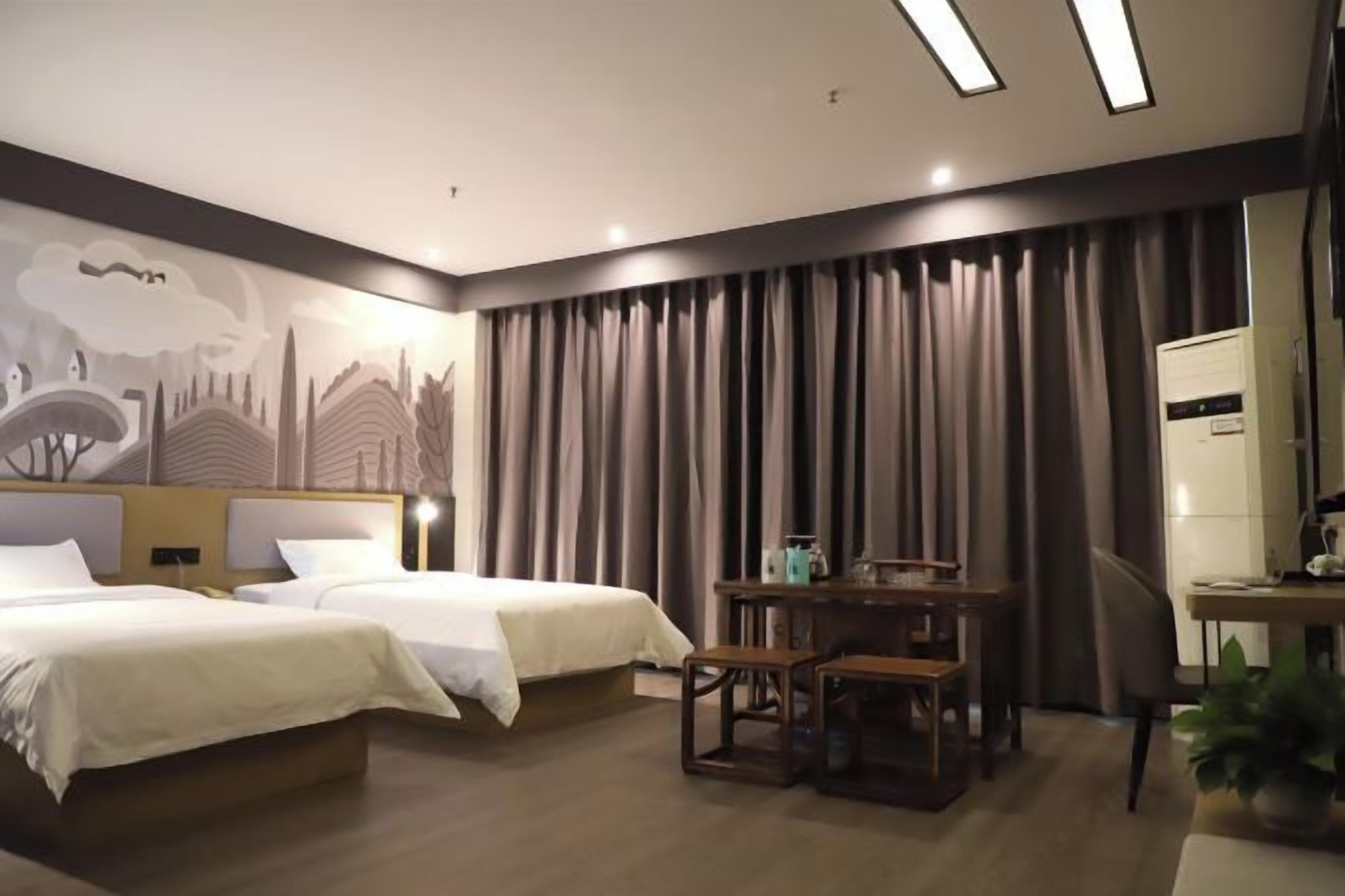 Quarto de hotel com duas camas, uma escrivaninha, uma cadeira e uma janela grande com cortinas.