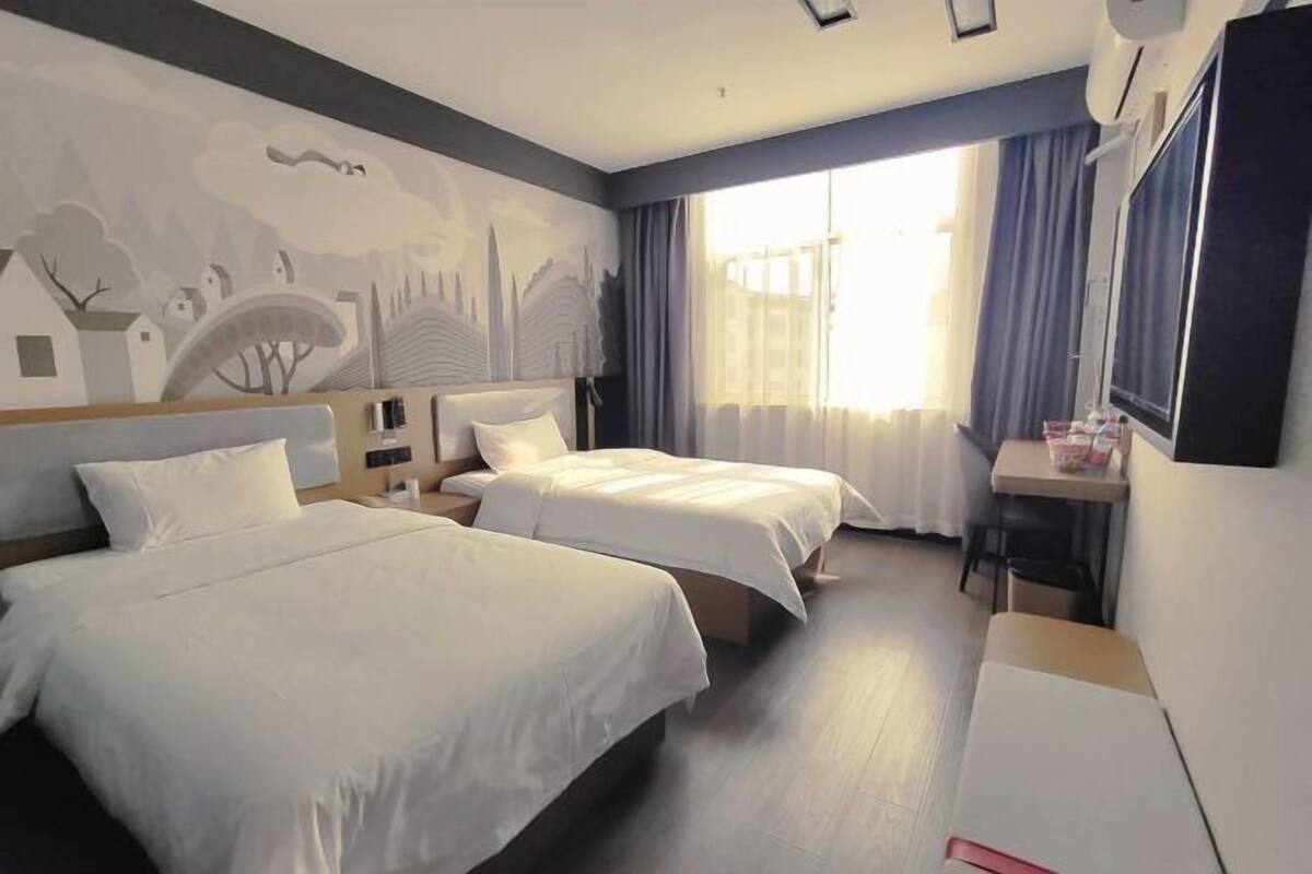 A hotel room with two beds, a desk, a chair, and a large window with curtains.