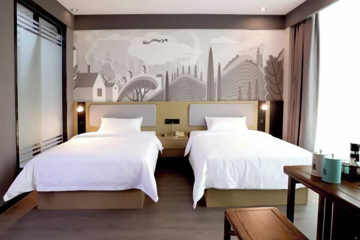 A hotel room with two beds, a mural on the wall, a bedside table with a lamp, and a window with curtains.