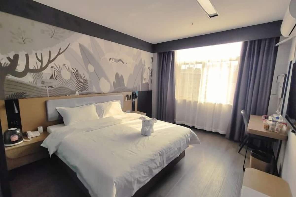 A hotel room with a large bed, a desk with a chair, a nightstand, and a wall mural.