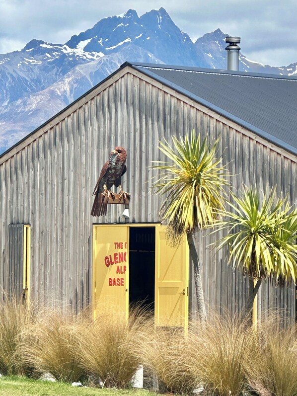 A rustic wooden building with a yellow door labeled "The Glen Alpine Base," a bird perched on the door, and a mountain range in the background.