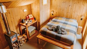 A cozy wooden room with a bed, a table with a lamp, and a wood stove. - The Great Glenorchy Alpine Base Camp (Glenorchy)