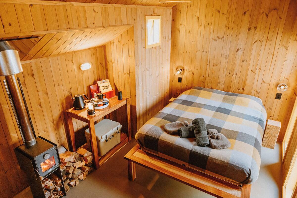 Une pièce chaleureuse en bois, avec un lit, une table et une lampe, ainsi qu’un poêle à bois.