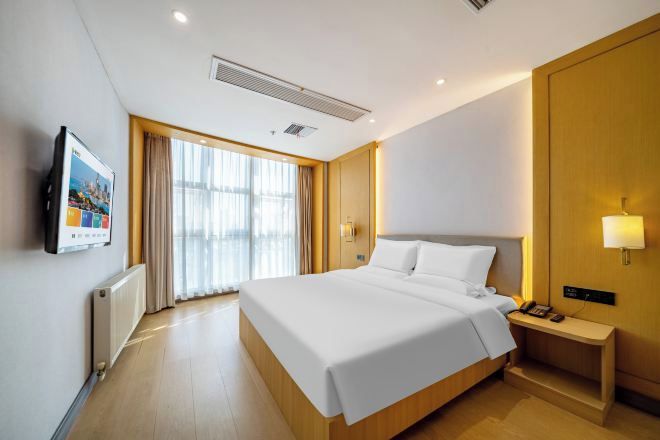 A modern hotel room with a large bed, a bedside table with a lamp, a TV mounted on the wall, and a large window with curtains.