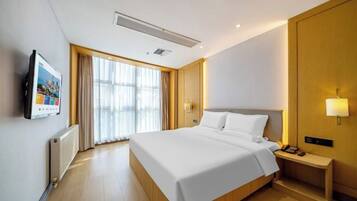 A modern hotel room with a large bed, a bedside table with a lamp, a TV mounted on the wall, and a large window with curtains.