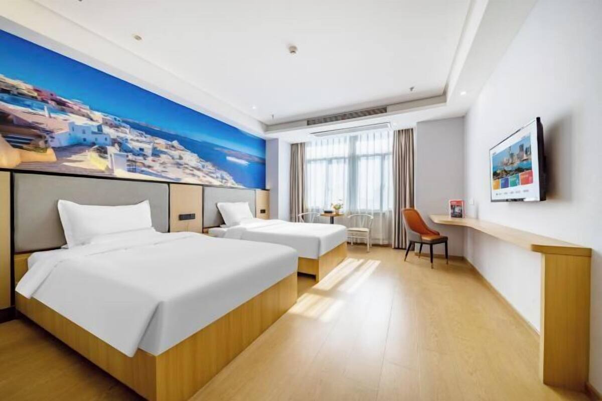 A hotel room with two beds, a desk, a chair, a TV, and a large painting of a coastal scene.