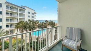 A balcony with a view of a pool and palm trees, a striped chair, and a multi-story building in the background. - BEACHFRONT RESORT, FAMILY FRIENDLY, TOP AMENITIES (Orange Beach)